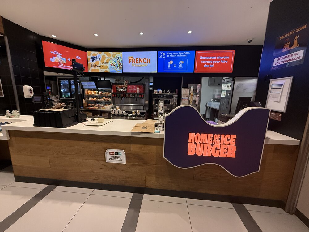 Zone de paiement bornes — vitrine à pâtisseries, écrans menu au-dessus du comptoir