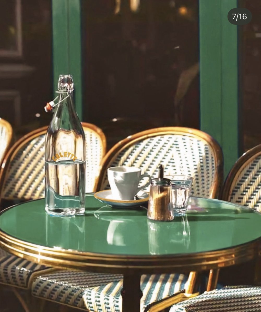 Terrasse Colette — table bistrot ronde plateau vert, chaises en rotin, bouteille d'eau logotypée (Lot 25)