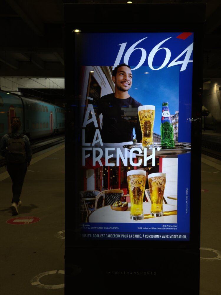 1664 « À la French » — serveur de bar posant avec pinte et bouteille verte, quai de gare la nuit