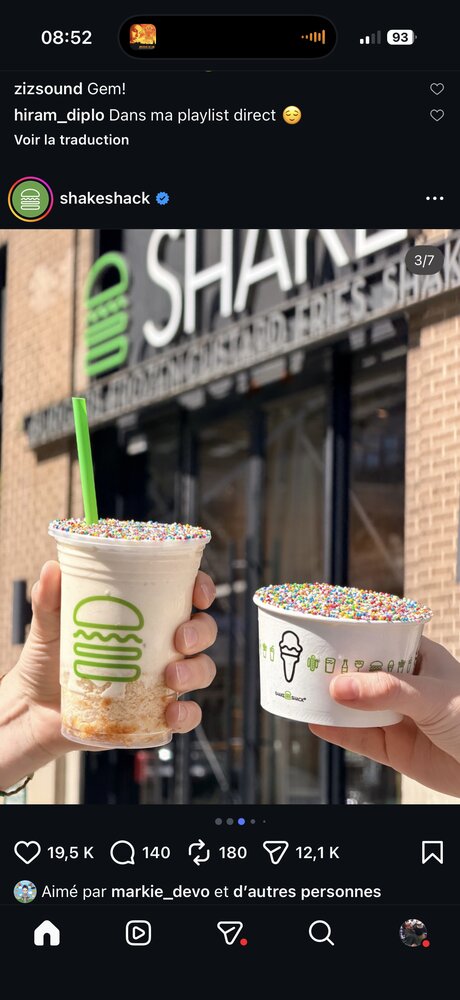 Deux mains tenant un milkshake et un sundae garnis de pépites colorées devant la façade Shake Shack