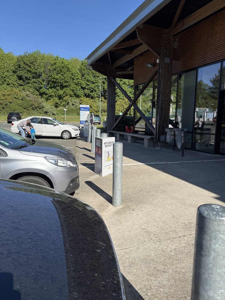 Vue parking et entrée Shell Café — structure bois en croix, signalétique « Prêt à boucler ton trajet »