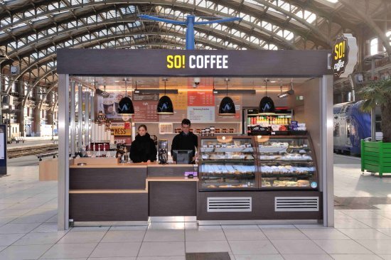 Kiosque « SO! Coffee » installé sous verrière de gare — comptoir bois foncé, vitrine viennoiseries, enseigne jaune/noire
